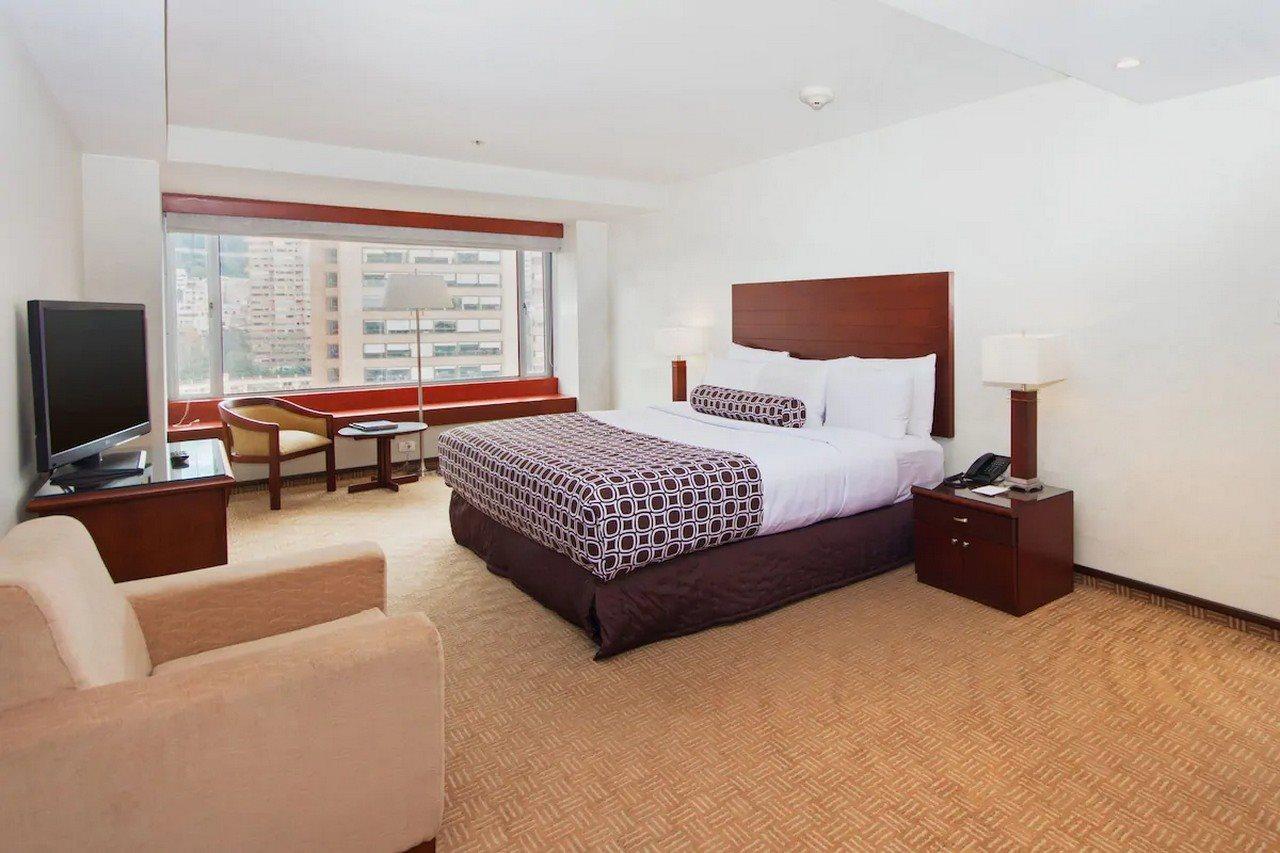 Torre Monserrate Bogota Exterior photo The image shows a contemporary hotel room. It features a large bed with white linens and a patterned bedspread, flanked by wooden nightstands with lamps. There is a comfortable armchair and a small table near a large window that allows natural light 