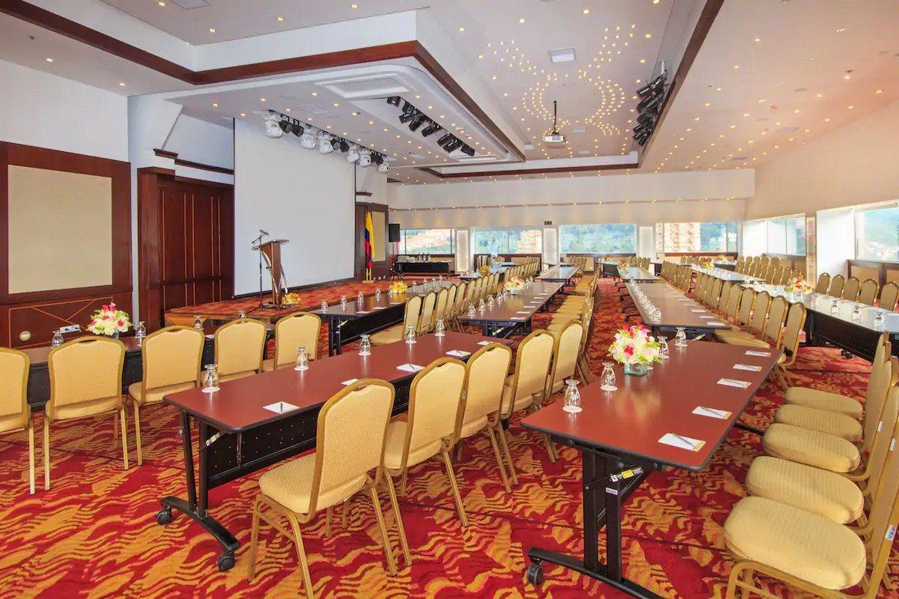 Torre Monserrate Bogota Exterior photo The photo shows a conference or meeting room set up for an event. The room features several long tables arranged in a U-shape, surrounded by yellow chairs. Each table has a notepad and pen placed on it. At the front of the room, there is a podium and