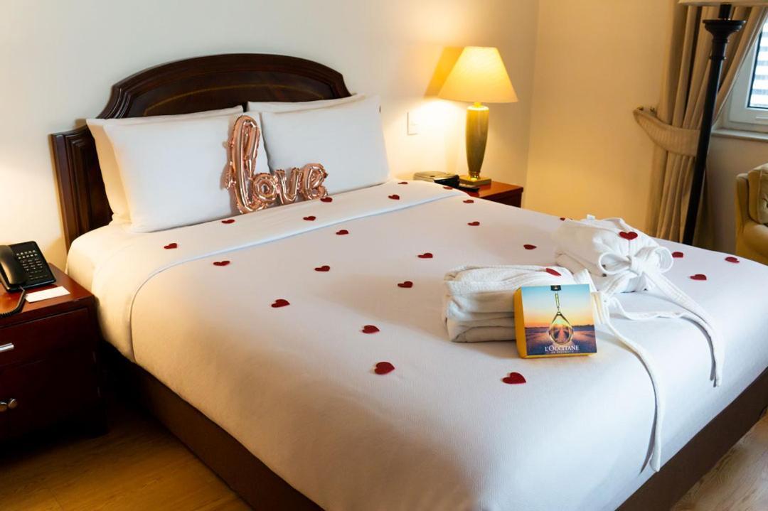 Torre Monserrate Bogota Exterior photo The photo shows a neatly arranged bed in a hotel room. The bed has white linens and is adorned with rose petals scattered across the top. There is a decorative pillow that says "love" in a metallic finish. Next to the bed, there is a lamp providing s