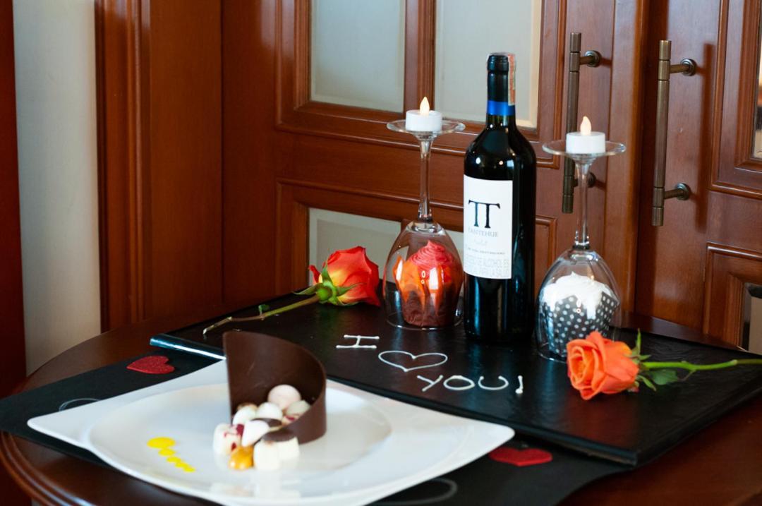 Torre Monserrate Bogota Exterior photo The photo features a romantic setup on a table. There is a plate with a chocolate dessert and some small candies or fruits. Surrounding the dessert are two roses, one orange and one red. In the background, there is a bottle of red wine labeled “IT” a