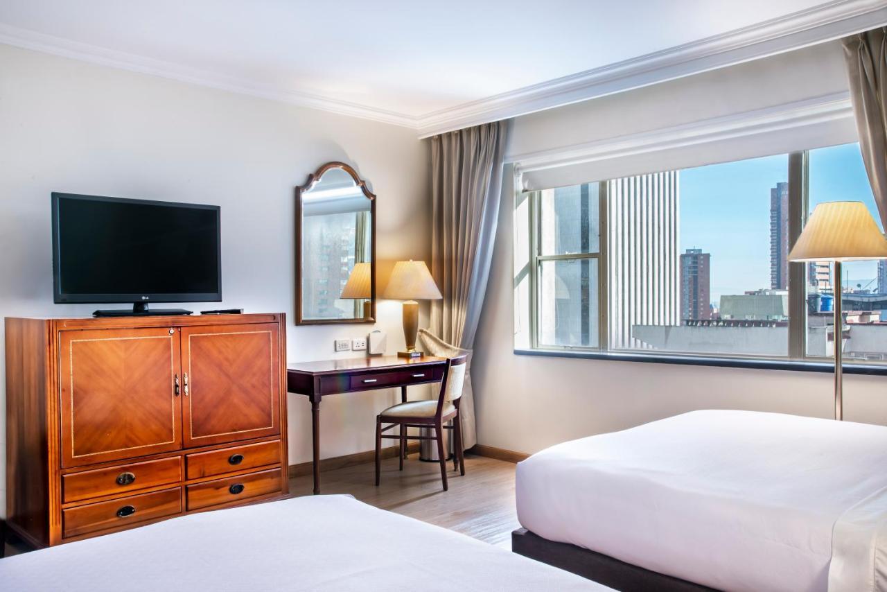Torre Monserrate Bogota Exterior photo The photo shows a hotel room with two single beds and modern decor. There's a flat-screen TV mounted on the wall, a wooden cabinet, and a desk with a chair positioned near a large window. The window offers a view of the outside, which includes tall b