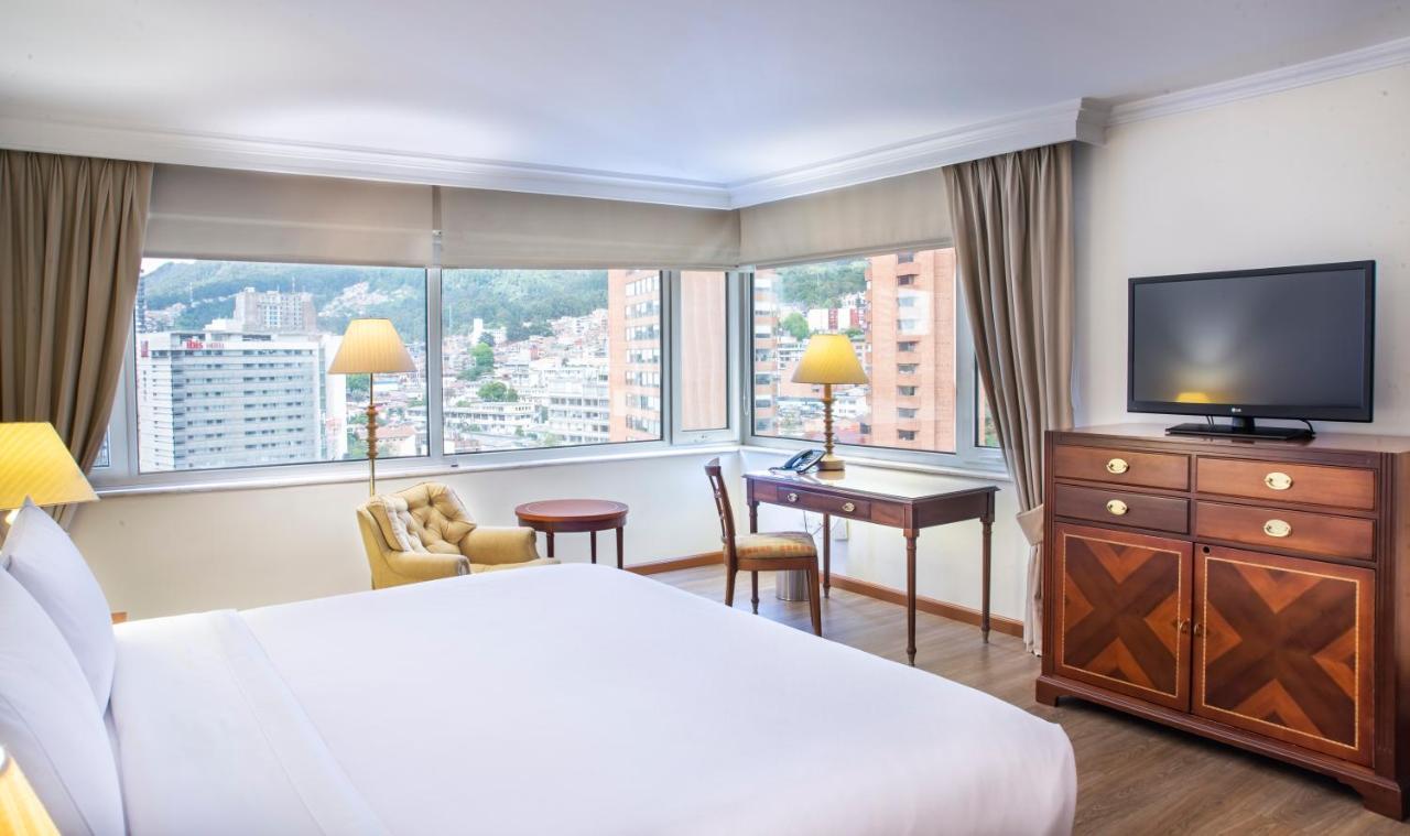 Torre Monserrate Bogota Exterior photo The photo shows a well-appointed hotel room featuring a large bed with a white comforter. There are two windows allowing natural light to flood the space, offering a view of the cityscape outside. In the room, there is a cozy armchair, a wooden desk 