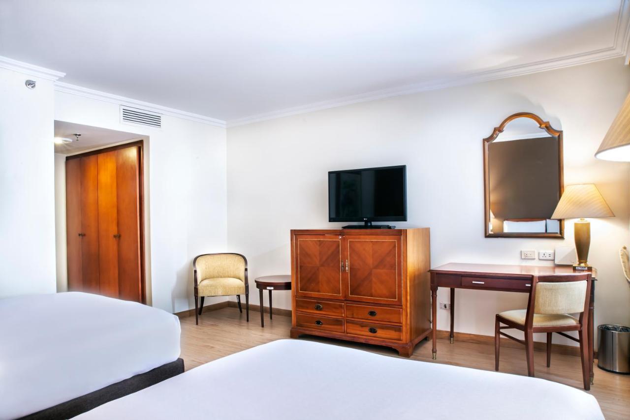 Torre Monserrate Bogota Exterior photo The photo shows a well-furnished hotel room featuring two beds with white bedding. There is a wooden dresser or cabinet in the center, which likely contains storage space. A flat-screen TV is mounted on the wall above the dresser. To the right, there