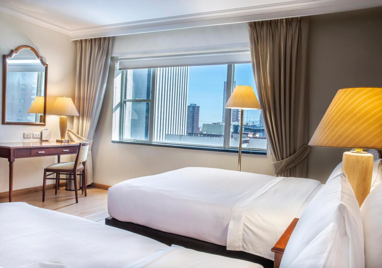 Torre Monserrate Bogota Exterior photo The photo shows a hotel room featuring two beds with white linens. There is a large window allowing natural light to fill the space, with a view of a cityscape visible outside. The room is decorated with light-colored curtains, and there are two lamp