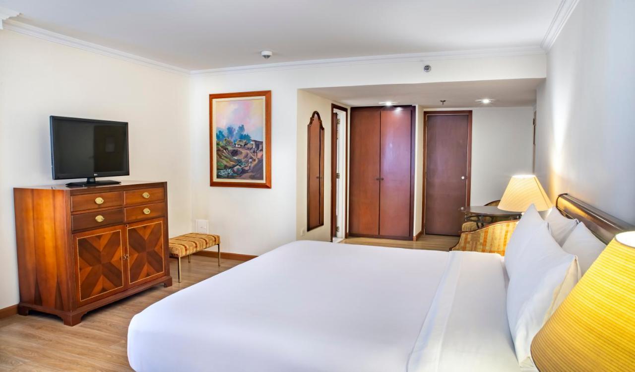 Torre Monserrate Bogota Exterior photo The photo shows a hotel room featuring a large, neatly made bed with white bedding. There's a wooden dresser with a television on top, and a chair nearby. A decorative painting hangs on the wall, adding a touch of color to the space. In the backgroun