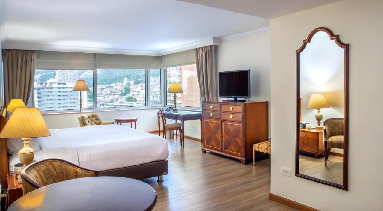Torre Monserrate Bogota Exterior photo The image shows a hotel room featuring a large window with natural light illuminating the space. There are two beds with white linens, a wooden dresser positioned against the wall, and a television mounted on a cabinet. A small desk and chair are in 