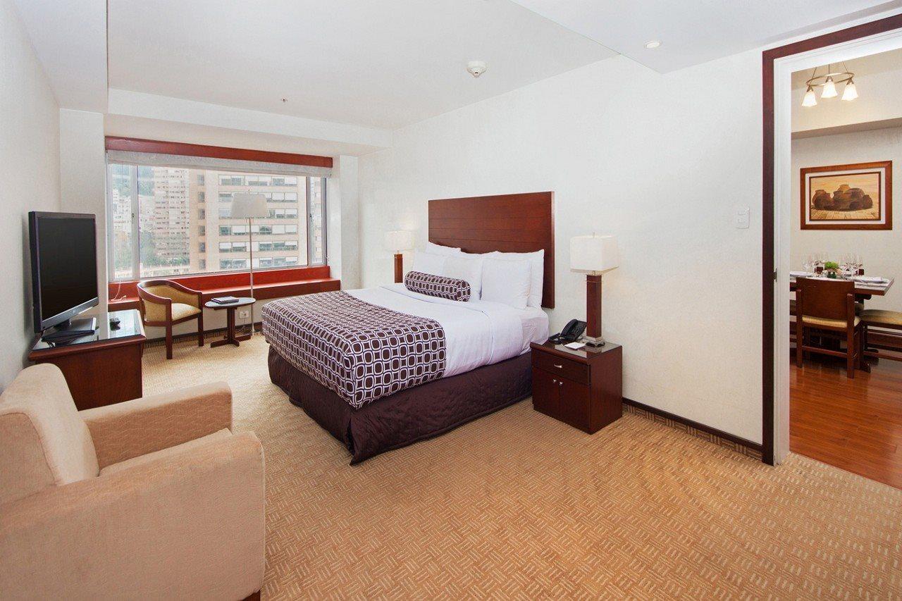 Torre Monserrate Bogota Exterior photo The photo shows a well-furnished hotel room. It features a large bed with a headboard, accompanied by a bedside table and a lamp. There is a seating area with a small couch and a couple of chairs, alongside a window that provides natural light and a 