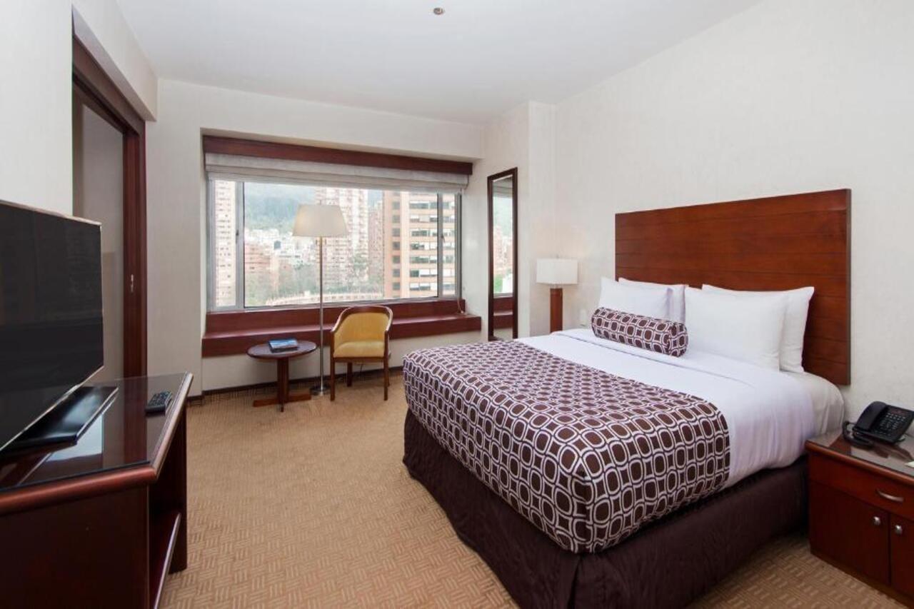 Torre Monserrate Bogota Exterior photo The photo shows a hotel room featuring a queen or king-sized bed with decorative bedding. There is a wooden headboard and a combination of white and patterned pillows. A large window provides natural light and offers a view of the city outside. Next 
