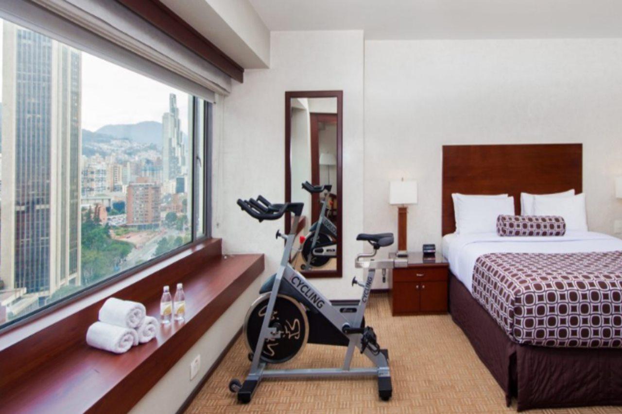 Torre Monserrate Bogota Exterior photo The photo shows a hotel room with modern decor. It features a bed with white and patterned bedding, a nightstand beside it with a lamp, and a full-length mirror mounted on the wall. In the foreground, a stationary exercise bike is positioned near a l