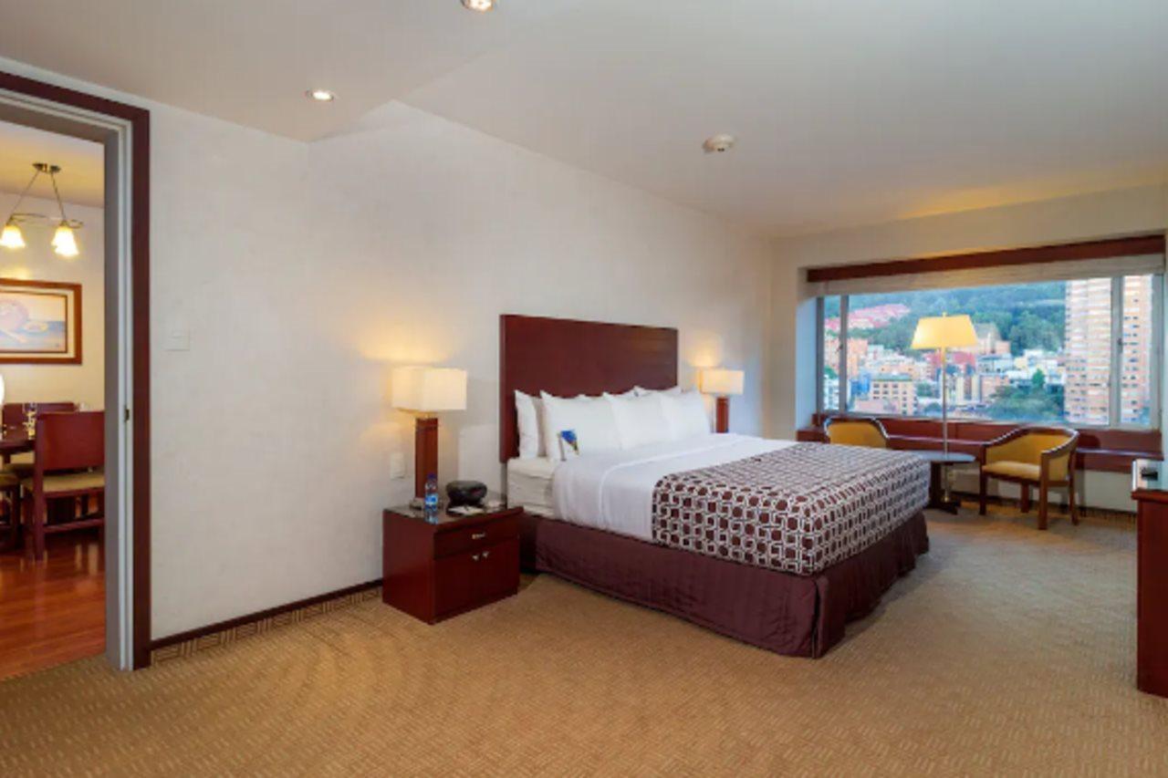 Torre Monserrate Bogota Exterior photo The photo shows a hotel room featuring a queen-sized bed with a patterned bedspread. There are two bedside lamps on either side of the bed, and a window with a view of the outside scenery, likely a cityscape or mountains. The walls are light in color