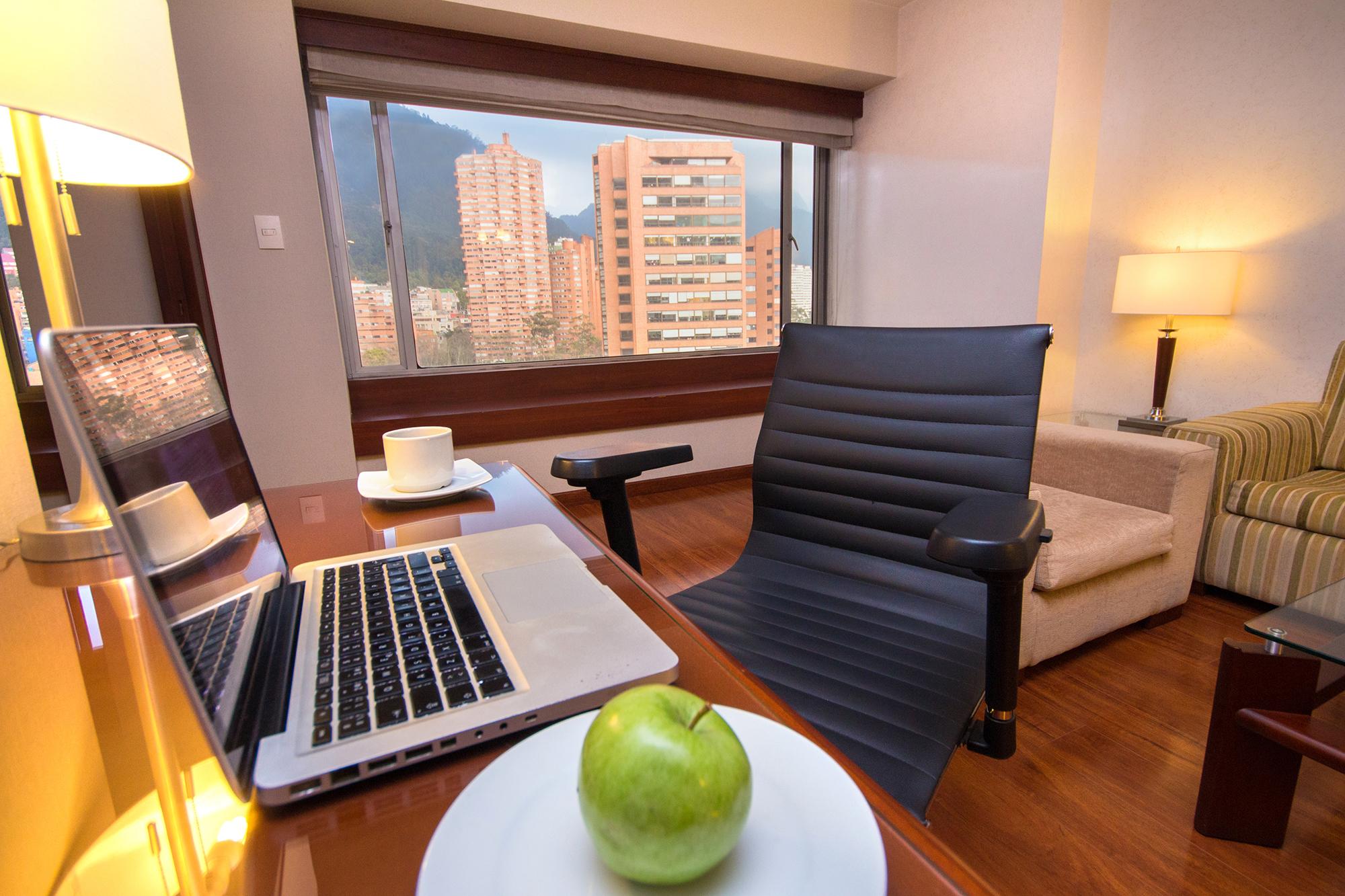 Torre Monserrate Bogota Exterior photo The photo shows a modern office space or hotel room with a view of buildings outside the window. In the foreground, there is a stylish desk with a laptop, a cup, and a green apple on a plate. A black ergonomic chair is positioned next to the desk. Th