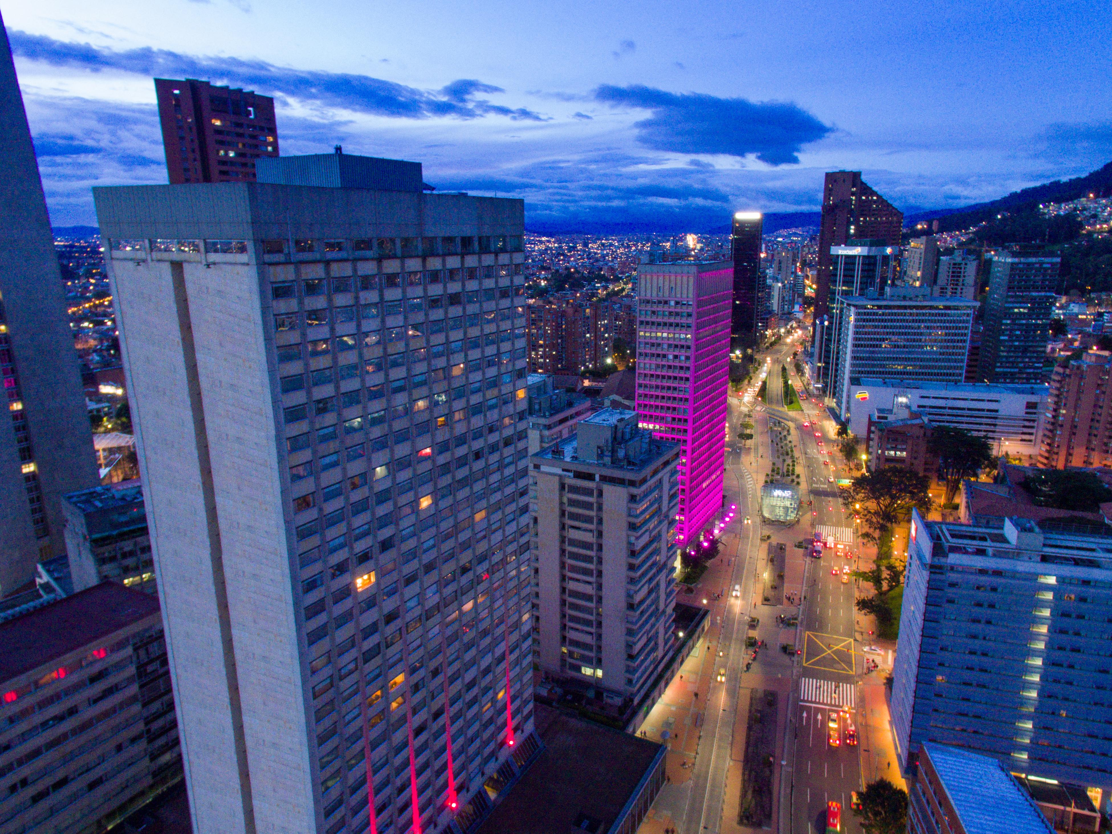 Torre Monserrate Bogota Exterior photo The photo shows an urban landscape during twilight. Skyscrapers and tall buildings are prominently featured, with one building illuminated in pink lights. The street below appears to be busy, with vehicles moving along it. In the background, addition