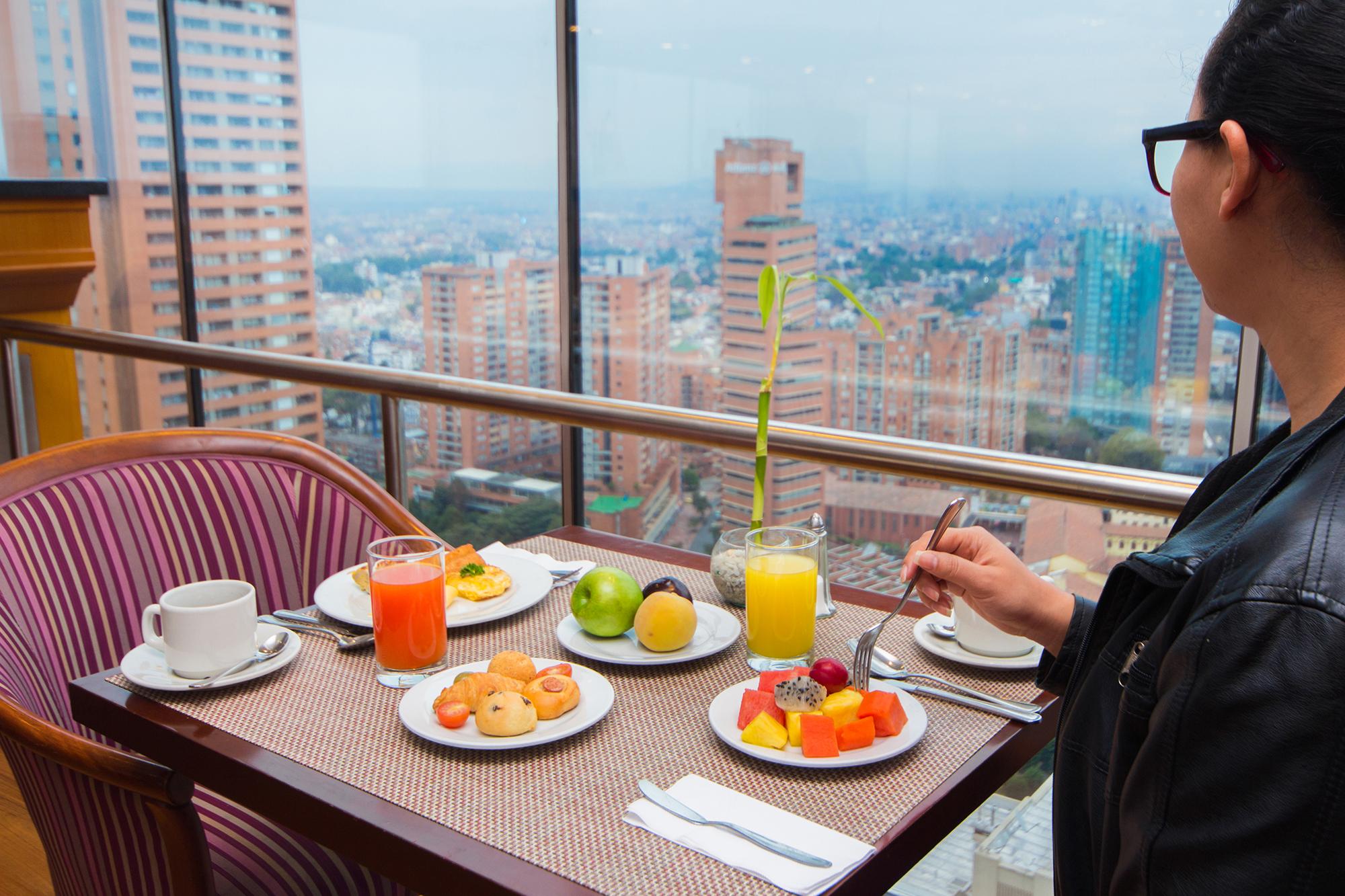 Torre Monserrate Bogota Exterior photo The photo depicts a breakfast scene at a table with a panoramic view of a city. The table is set with several plates: one with a colorful assortment of fresh fruit, another with small pastries or donuts, and a third with a beverage. There are two dri