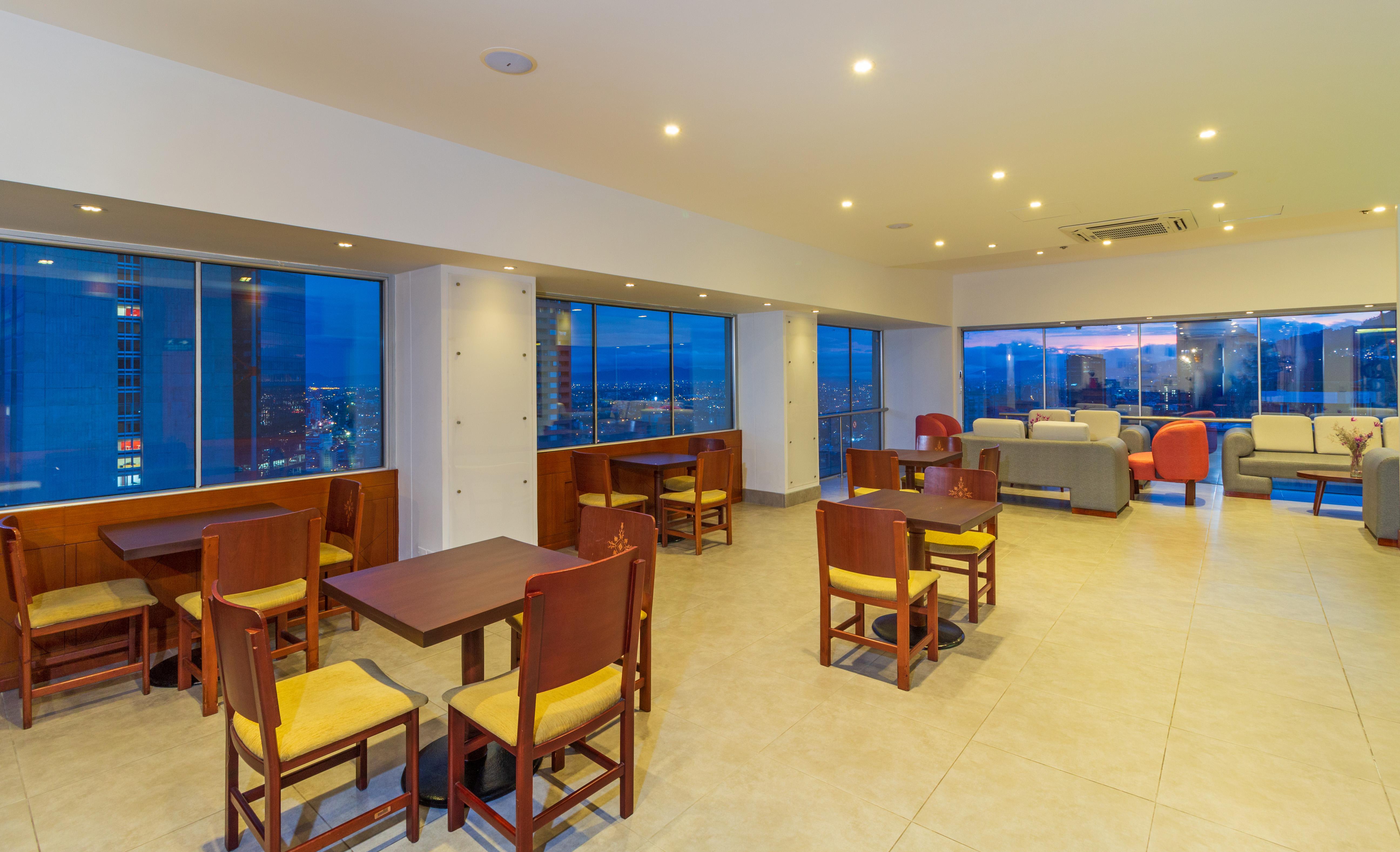 Torre Monserrate Bogota Exterior photo The photo shows a spacious, modern interior of a café or lounge area with large windows, providing a view of the city. The flooring is tiled, and the lighting is warm, creating a welcoming atmosphere. There are several wooden tables with yellow chair