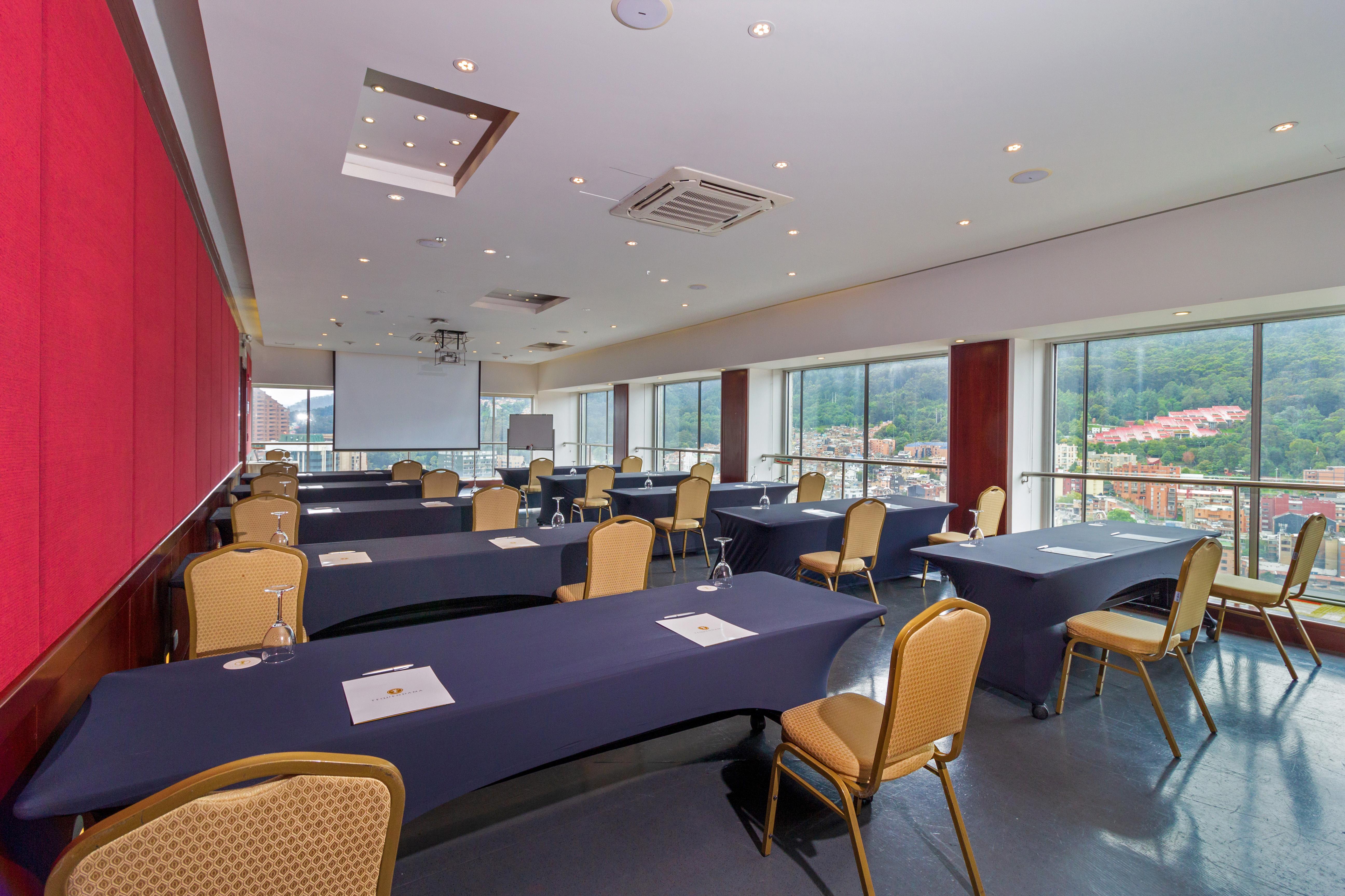 Torre Monserrate Bogota Exterior photo The photo shows a conference room designed for meetings or presentations. It features several tables arranged in a rectangle, each covered with dark blue tablecloths. There are chairs with golden fabric surrounding the tables. The room has large wind
