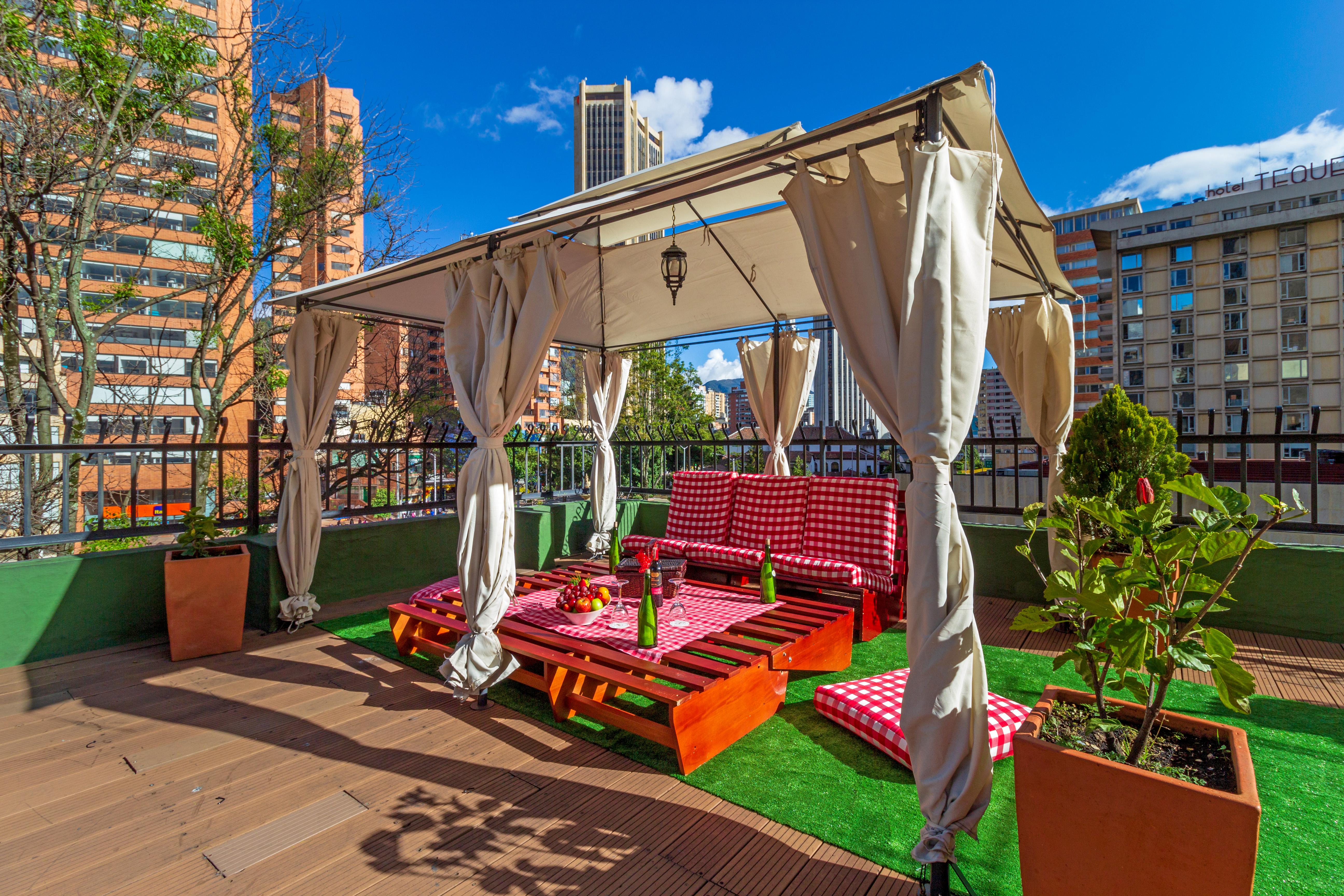Torre Monserrate Bogota Exterior photo The photo showcases a cozy outdoor lounging area, likely on a rooftop or terrace. It features a canopy with draped curtains, providing shade and an inviting atmosphere. Beneath the canopy, there are wooden platforms arranged with cushions in a red an