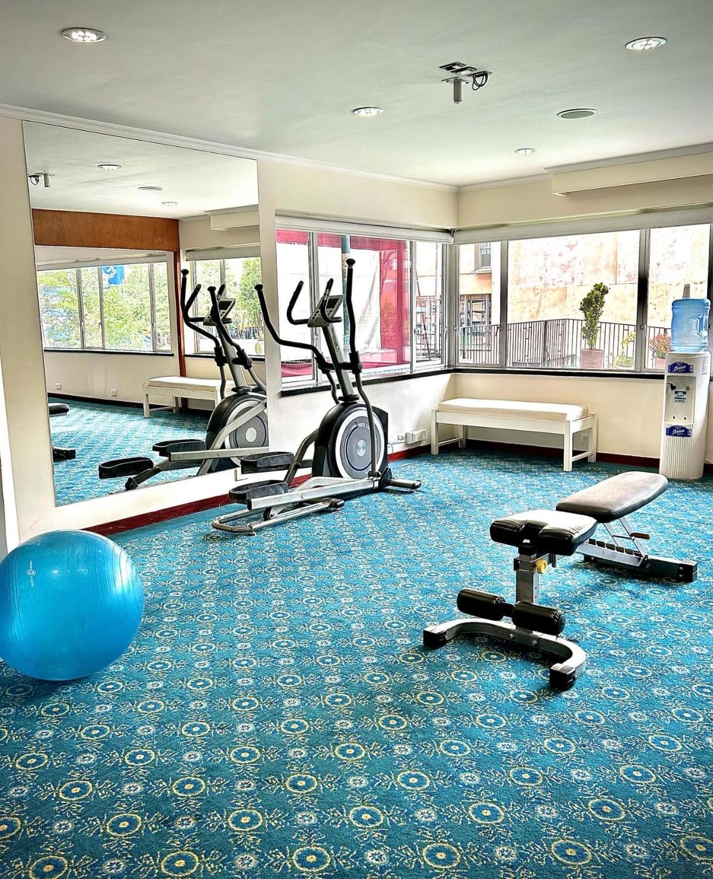 Torre Monserrate Bogota Exterior photo The photo shows a small gym or fitness area. In the space, there are two elliptical machines positioned next to large windows that allow natural light to enter. There's also a workout bench and a blue exercise ball on the floor, which features a patt