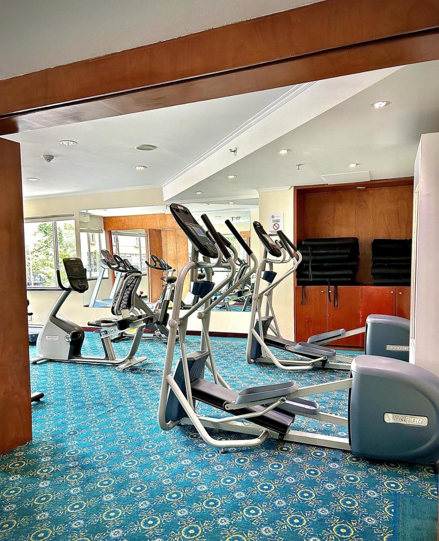 Torre Monserrate Bogota Exterior photo The photo shows a fitness room equipped with several exercise machines. There are two elliptical machines and a couple of stationary bikes arranged in the space. The floor is covered with a blue carpet that has a patterned design. Large windows allow